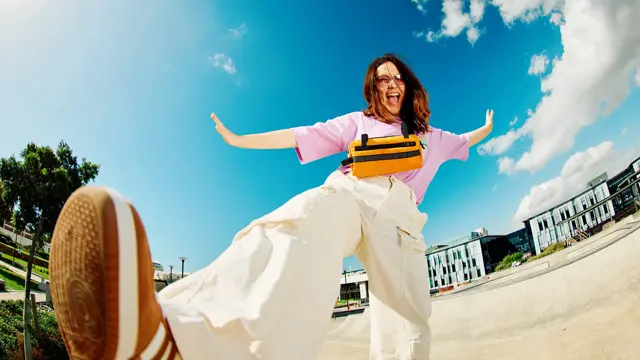 Dame med brune sko, hvit bukse og rosa topp smiler og sparker til kamera som ligger på bakken med blå himmel i bakgrunnen