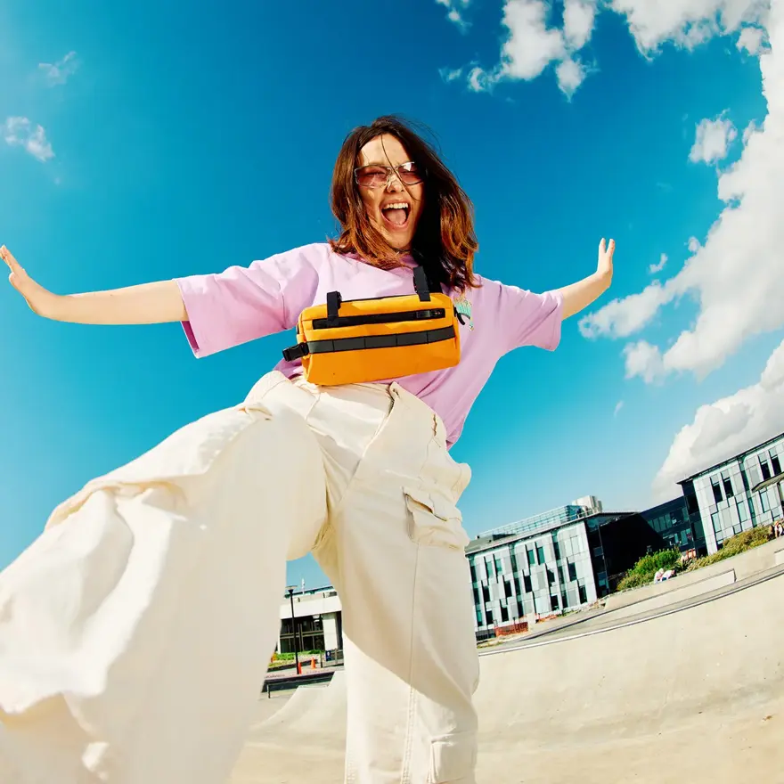 Dame med brune sko, hvit bukse og rosa topp smiler og sparker til kamera som ligger på bakken med blå himmel i bakgrunnen