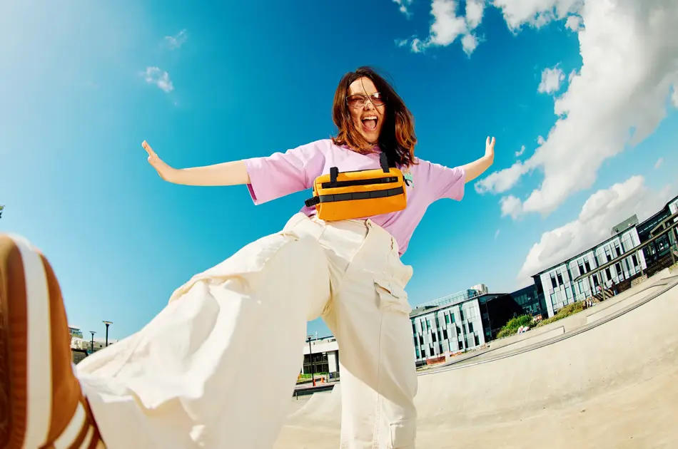 Dame med brune sko, hvit bukse og rosa topp smiler og sparker til kamera som ligger på bakken med blå himmel i bakgrunnen