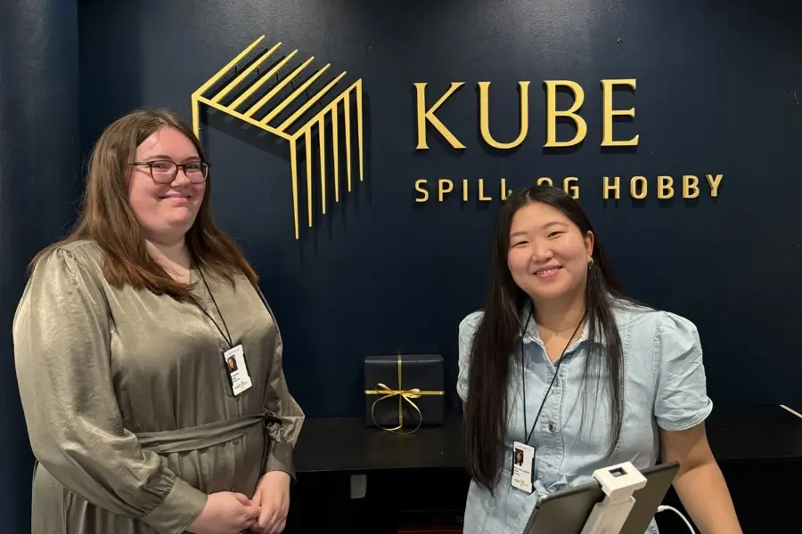 To damer bak en kassedisk med logoen KUBE spill og hpbby