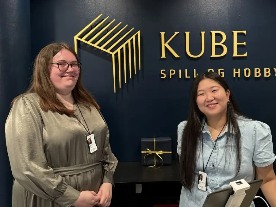 To damer bak en kassedisk med logoen KUBE spill og hpbby