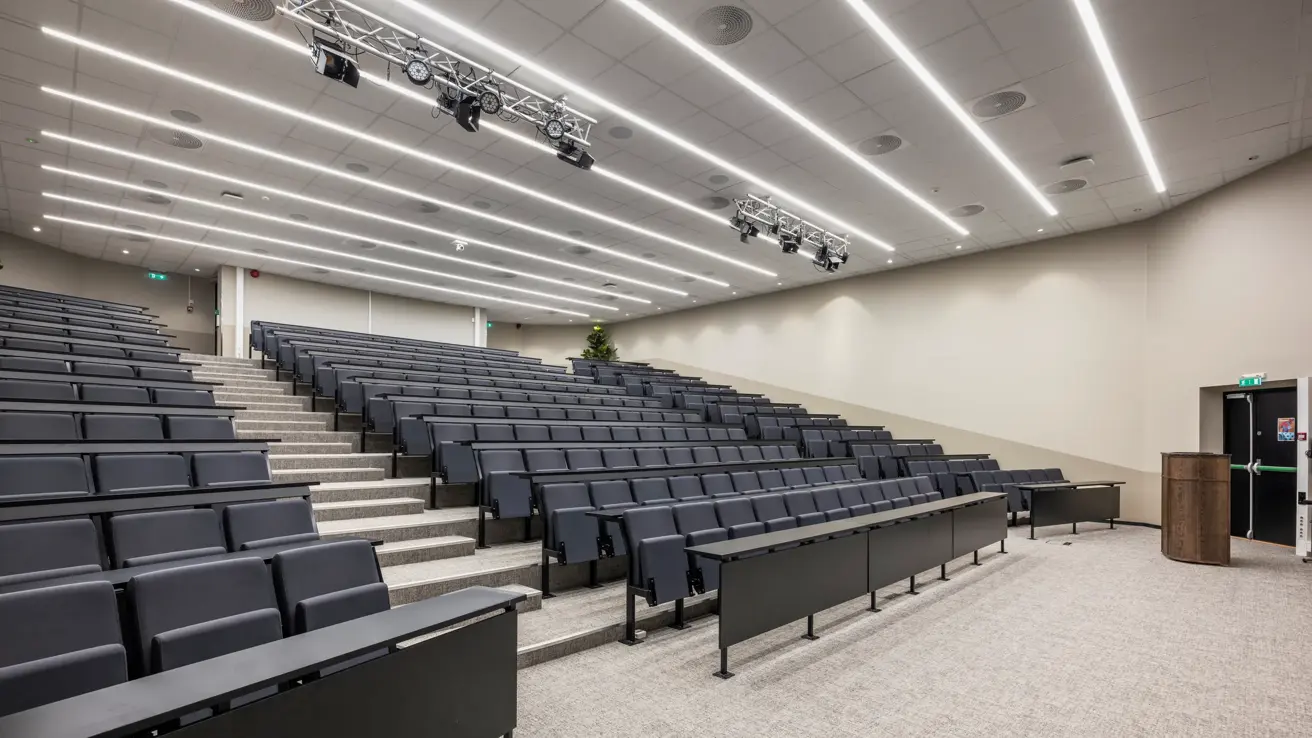 Auditorium med blå stoler og svarte bord, talerstol foran ved scenen.