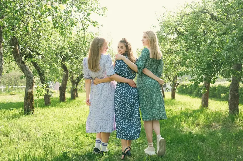 Tre jenter med blomsterkjoler holder rundt hverandre i en hage sett bakfra 