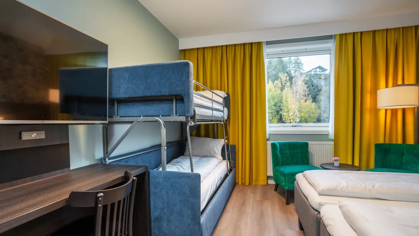 Familierom med twin senger, køyesenger, skrivebord, stol, TV på veggen, grønne lenestoler og gule gardiner.