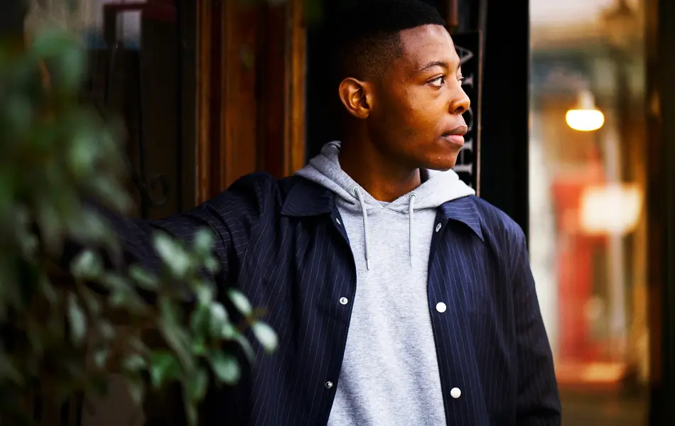 Mann poserer med blazer og grå hettegenser, grønne planter i forgrunn og trevegg i bakgrunnen