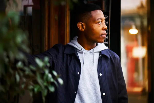 Mann poserer med blazer og grå hettegenser, grønne planter i forgrunn og trevegg i bakgrunnen