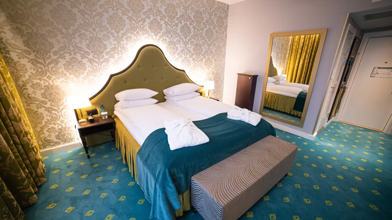 Juniorsuite med dobbeltseng, morgenkåper, gul sengegavl, mønstrete, lys tapet og grønt vegg-til-vegg-teppe med bristol logoer.