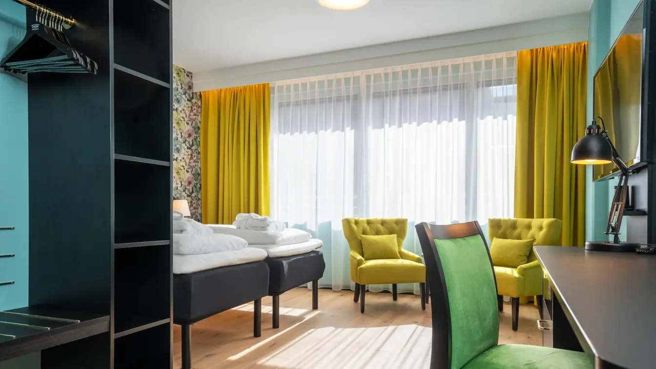 Åpen garderobe med kleshengere. Separate senger, sittegruppe med gule stoler, skrivebord med grønn stol. Gule gardiner og store vinduer i superior rom på Thon Hotel Cecil.