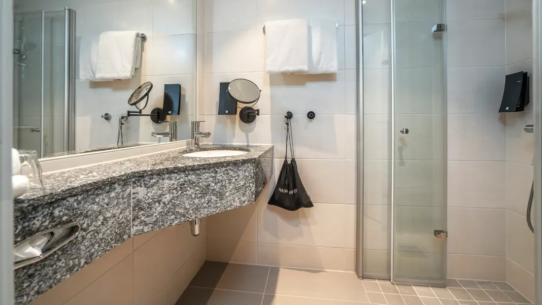 Bad på twinrom med servant, dusjhjørne, håndkleoppheng på vegg, stort speil på vegg og mindre speil på vegg, flisebelagt gulv og vegger. Såpedispenser ved servant og i dusj.