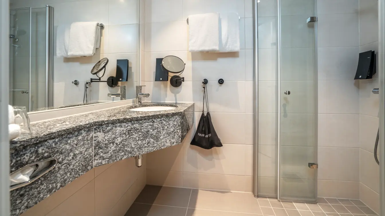 Bad på twinrom med servant, dusjhjørne, håndkleoppheng på vegg, stort speil på vegg og mindre speil på vegg, flisebelagt gulv og vegger. Såpedispenser ved servant og i dusj.