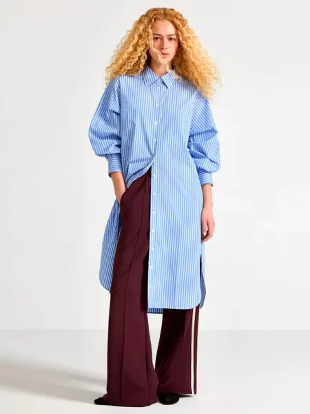 Damemodell med lang blåstripete skjortekjole, burgunder bukse og sorte sko.
