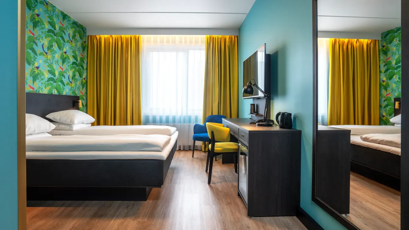 Grønn tapetvegg med mønster, gule gardiner, dobbeltseng, parkettgulv, blå stol, skrivebord, vindu, gul stol, speil