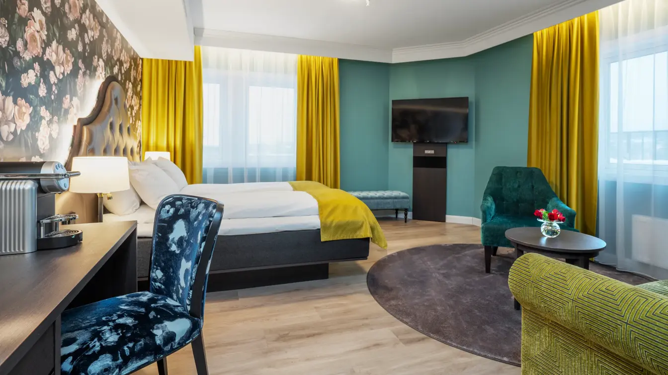 Mørk tapet med store lyserosa roser. Vindu med gule gardiner fra tak til gulv. Dobbeltseng med gult sengeteppe. Mørkt skrivebord med kaffemaskin og mønstrete stol. Turkis vegg og veggehengt TV. I superior rom på Thon Hotel Høyers i Skien.