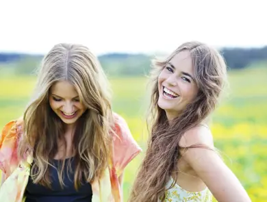 To smilende damer med oransje og gul topp med gul blomstereng i bakgrunnen 