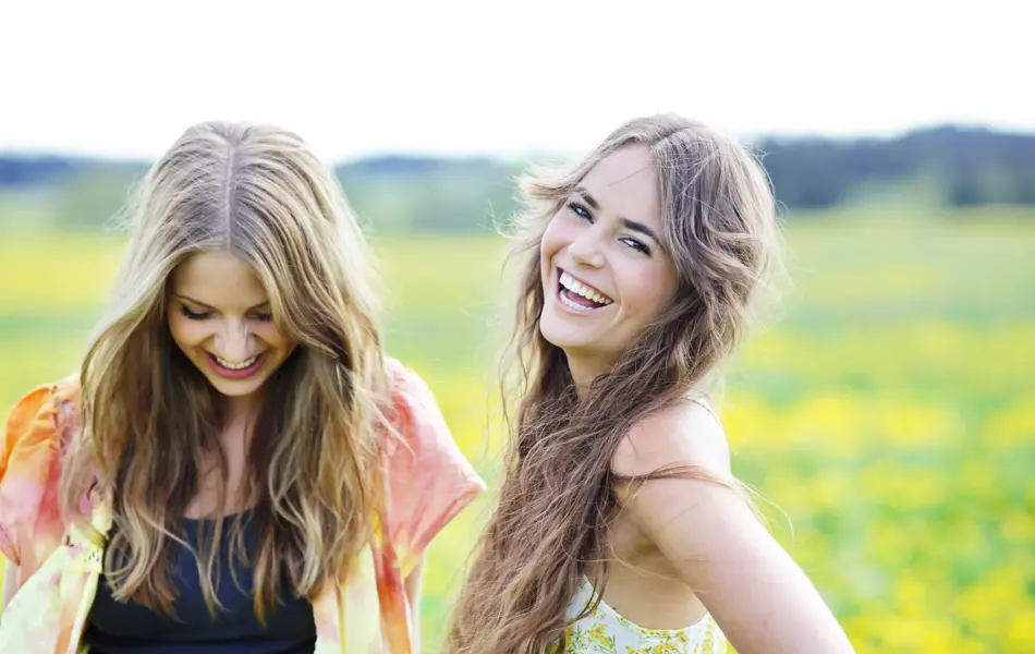 To smilende damer med oransje og gul topp med gul blomstereng i bakgrunnen 