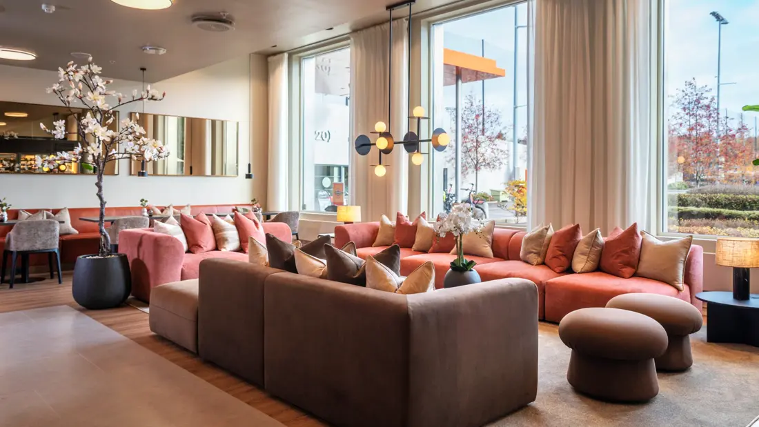 Lobbyområde med flere sittegrupper, store sofaer, små bord, duse farger, store vinduer og lange hvite gardiner
