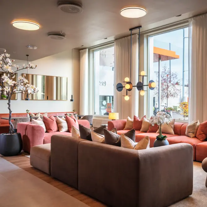 Lobbyområde med flere sittegrupper, store sofaer, små bord, duse farger, store vinduer og lange hvite gardiner