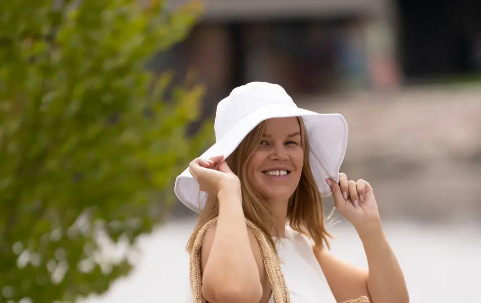 Dame står ute med hvit hatt, hvit kjole og en stråveske over skulderen