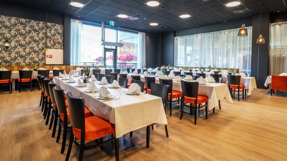 Restaurant dekket opp med tre langbord, med hvite duker, svarte stoler med oransje putetrekk, mønstrete tapet og store vinduer.