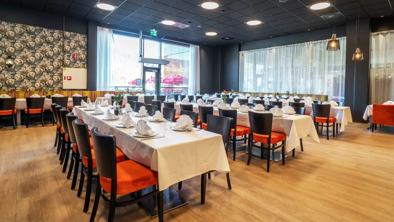 Restaurant dekket opp med tre langbord, med hvite duker, svarte stoler med oransje putetrekk, mønstrete tapet og store vinduer.