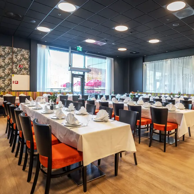 Restaurant dekket opp med tre langbord, med hvite duker, svarte stoler med oransje putetrekk, mønstrete tapet og store vinduer.