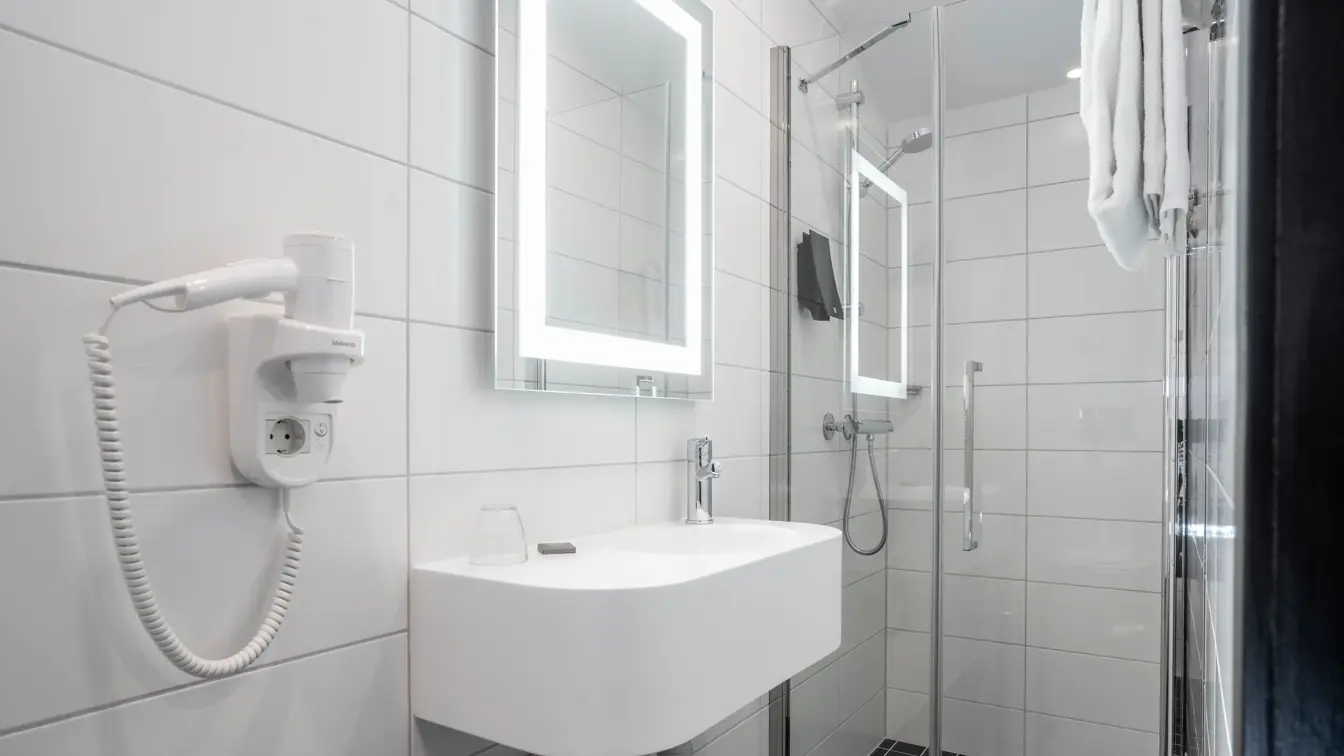 Veggfestet hårføn, servant, speil med innebygget lys, hvit kakel og dusj i baderom på Thon Hotel Astoria