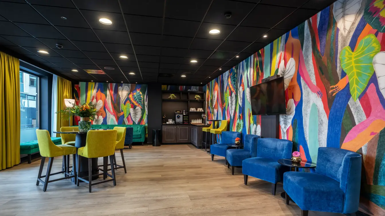 Lobbyområde med blå lenestoler langs veggen, rundt stort bord med gule høye stoler rundt. Fargerik tapet på veggene og gule gardiner.