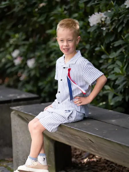 Liten gutt sitter ute på en benk med blå og hvitstripete skjorte og shorts, 17. mai sløyfe og beige tøysko.