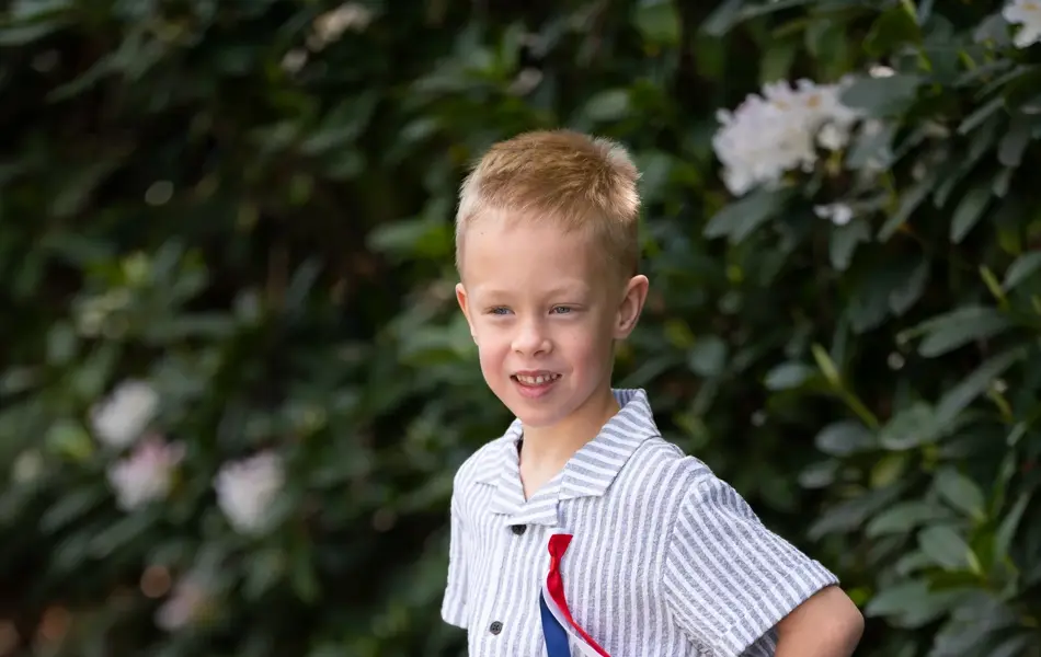 Liten gutt sitter ute på en benk med blå og hvitstripete skjorte og shorts, 17. mai sløyfe og beige tøysko.