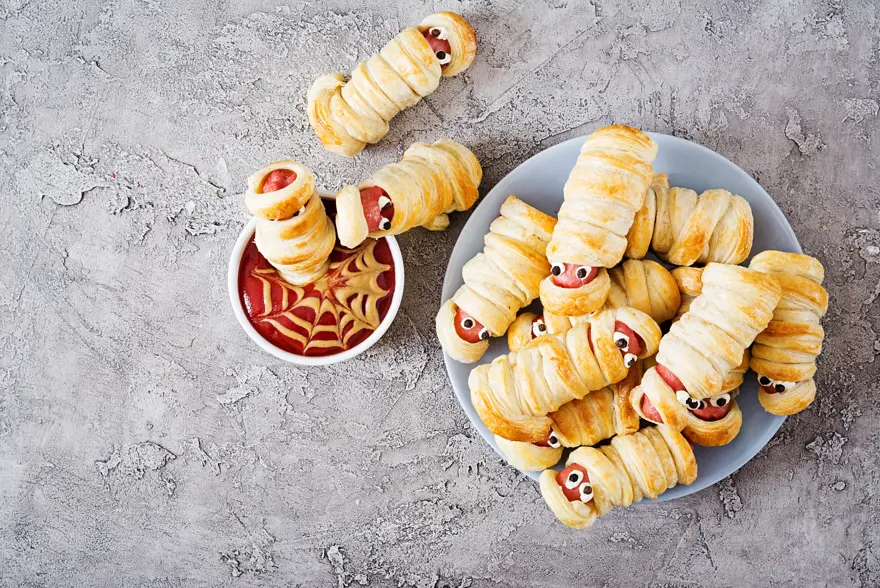 Scary sausage mummies in dough with funny eyes on table. Funny decoration. Halloween food. Top view. Flat lay