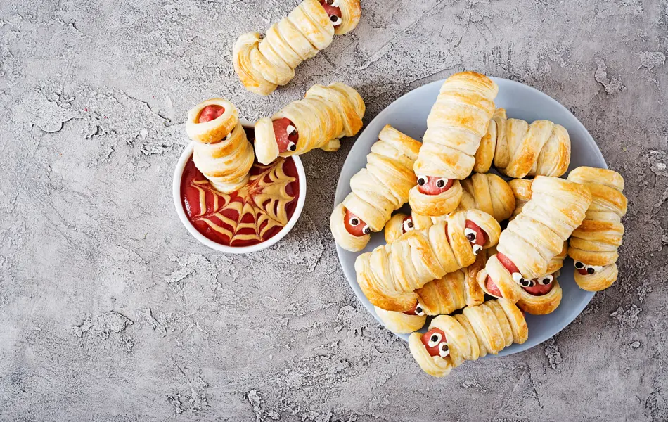 Scary sausage mummies in dough with funny eyes on table. Funny decoration. Halloween food. Top view. Flat lay