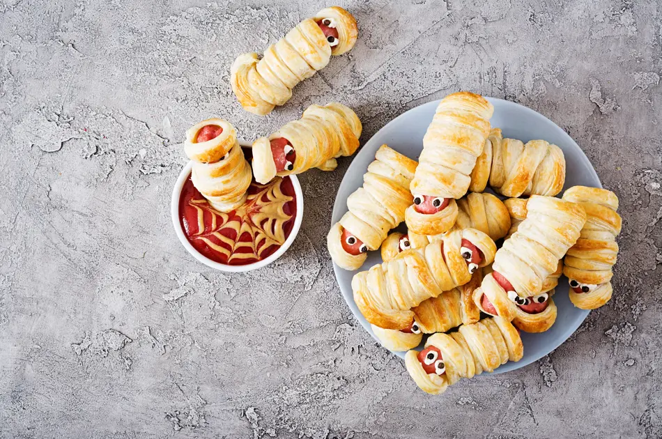 Scary sausage mummies in dough with funny eyes on table. Funny decoration. Halloween food. Top view. Flat lay