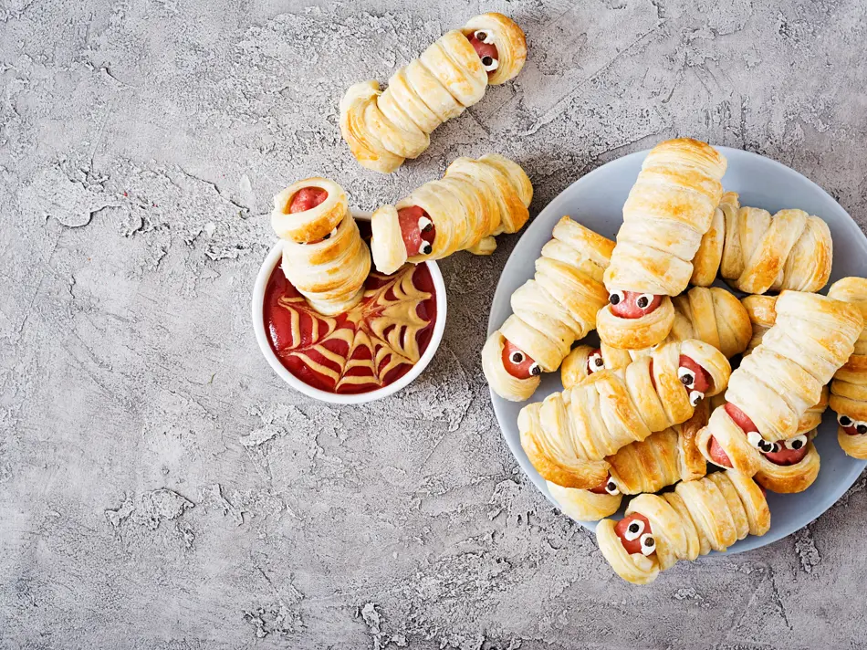 Scary sausage mummies in dough with funny eyes on table. Funny decoration. Halloween food. Top view. Flat lay