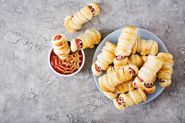 Scary sausage mummies in dough with funny eyes on table. Funny decoration. Halloween food. Top view. Flat lay