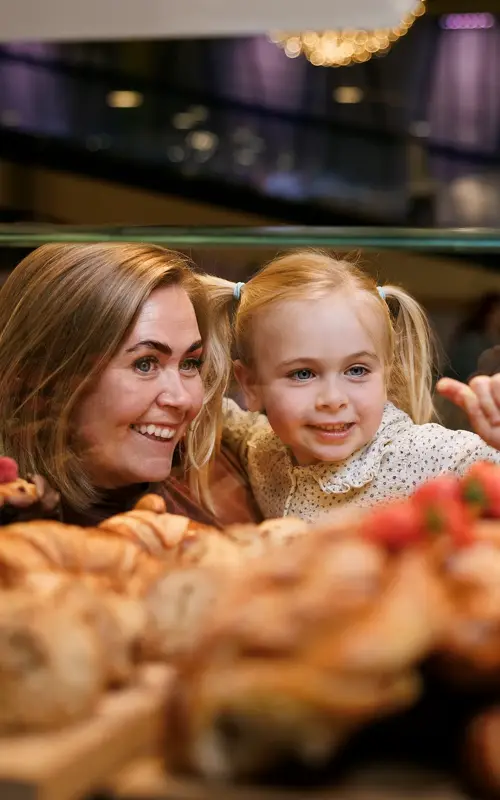 En kvinne og en liten jente står og ser inn gjennom glasset på bakverk som ligger der