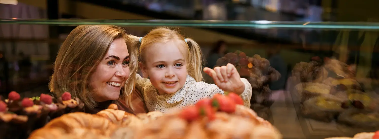 En kvinne og en liten jente står og ser inn gjennom glasset på bakverk som ligger der