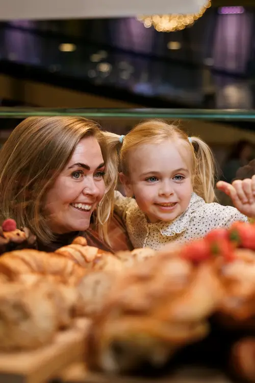 En kvinne og en liten jente står og ser inn gjennom glasset på bakverk som ligger der