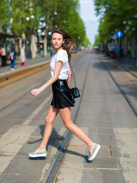 Kvinne med langt hår går over veien i svart miniskjørt i skinn, hvit t-skjorte og svart veske.