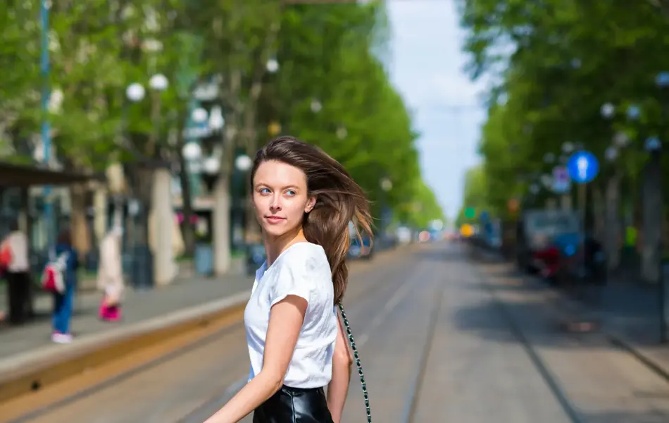 Kvinne med langt hår går over veien i svart miniskjørt i skinn, hvit t-skjorte og svart veske.