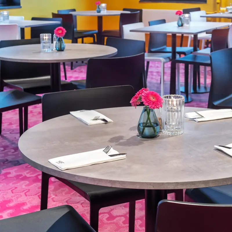 Frokostsal, rosa teppe, svarte runde spisebord, svarte stoler, vindu, gule vegger, gule gardiner. 