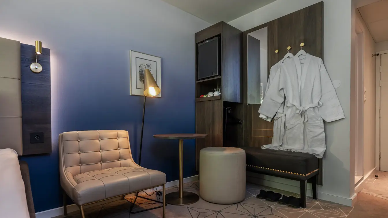 Superior rom med diverse møbler. Lenestol, lite bord, puff, stålampe, minibar og to morgenkåper som henger på veggen