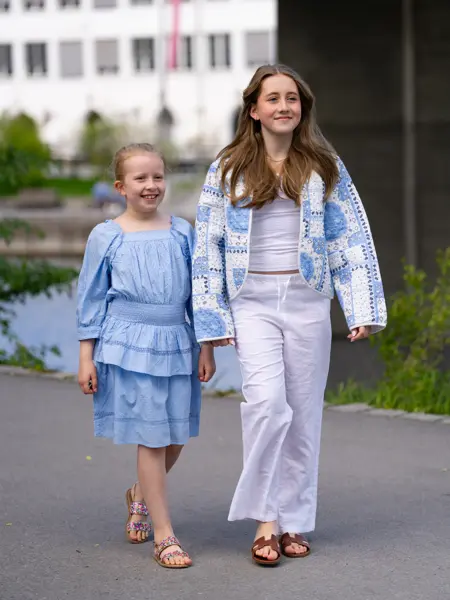 Jente i blå kjole og sandaler, større jente i hvit bukse og topp, brune sandaler og mønstret blå jakke.