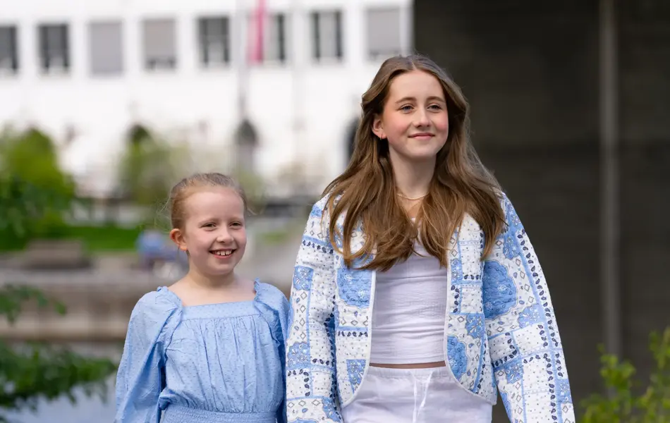 Jente i blå kjole og sandaler, større jente i hvit bukse og topp, brune sandaler og mønstret blå jakke.
