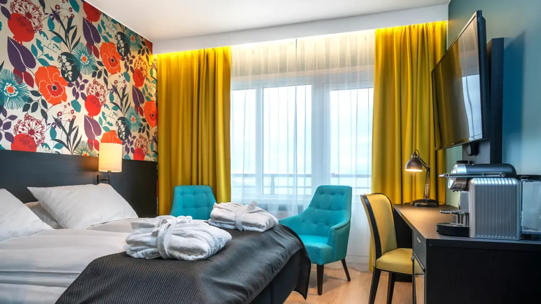 Blomstrete tapet, et stort vindu fra gulv til tak, gule gardiner, to blå stoler, et lite bord, en dobbeltseng, svart skrivebord, gul stol og en flatskjerm hengende på veggen. 