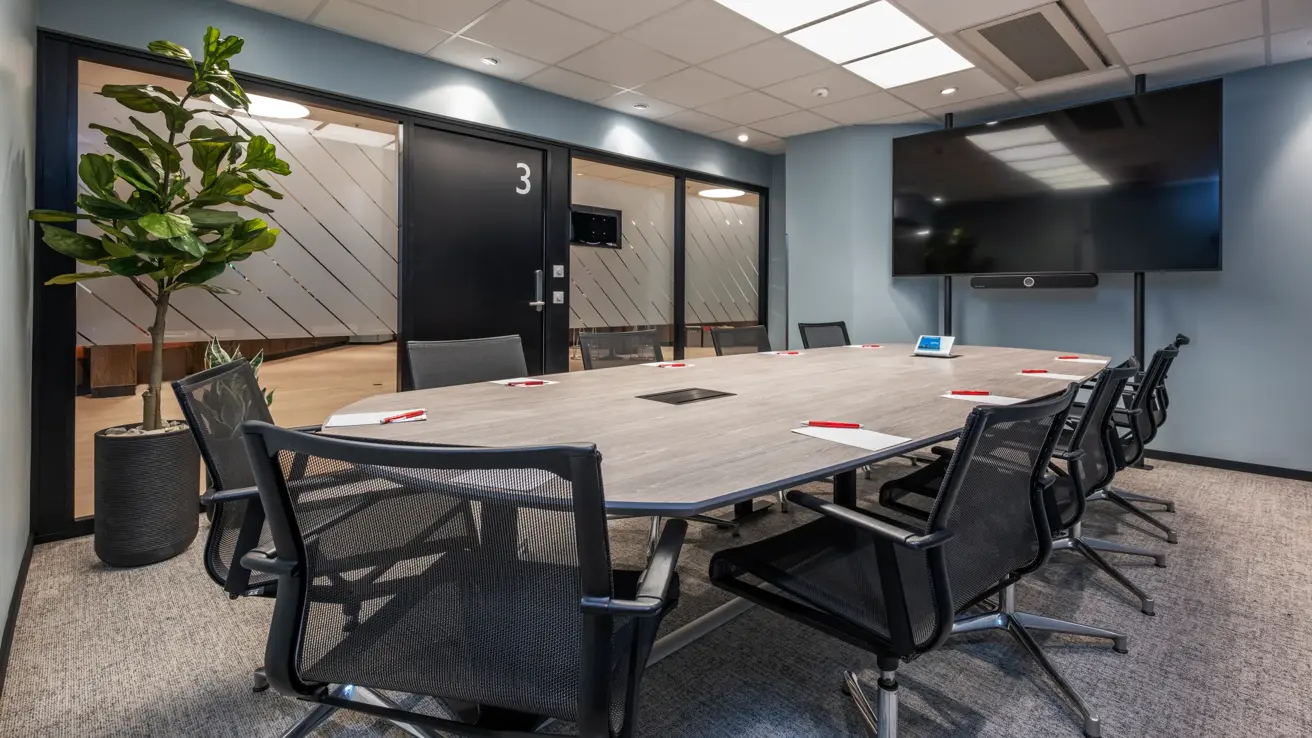 Møterom med styrebords oppsett til 10 personer. Grønn stor plante i hjørnet, stor skjerm, vinduer langs den ene langsiden ut i gang. Penn og papir på bordet.