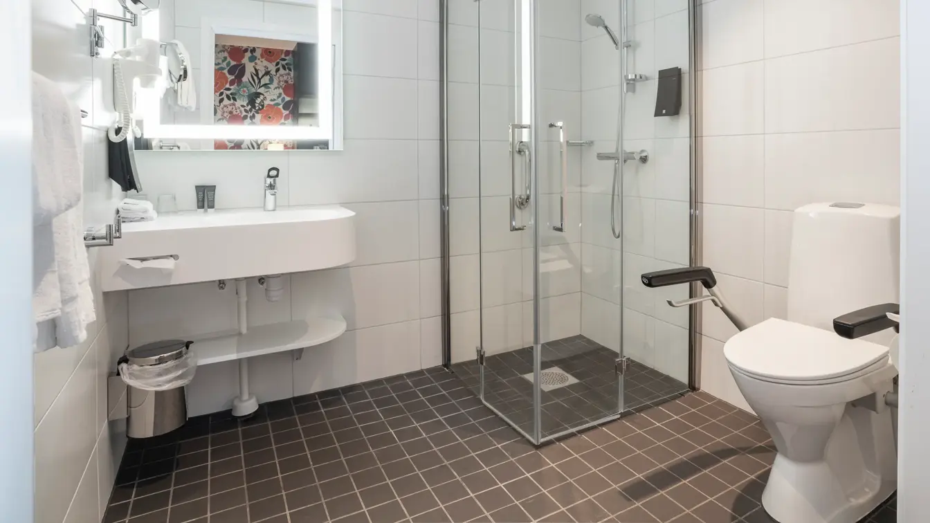 Svarte gulvfliser med hvite veggfliser, hvit porselens toalett med armlener, glassdusj kabinett og en hvit porselens vask.