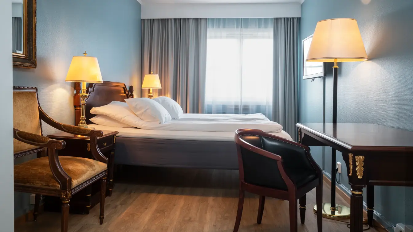 Dobbeltseng, to lamper, skrivebord, to stoler, vindu, grå gardiner, parkettgulv, turkise vegger. 