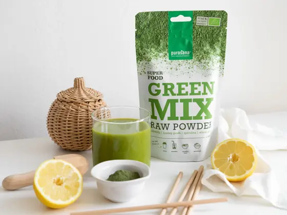 En pose med Green Mix fra Purasana står ved siden av et glass med en grønn væske og to halve sitroner.