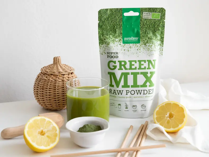 En pose med Green Mix fra Purasana står ved siden av et glass med en grønn væske og to halve sitroner.