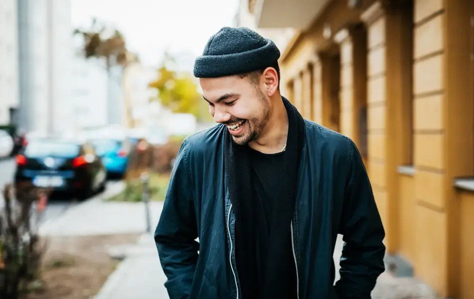 Smilende ung mann i urbane omgivelser, iført et fasjonabelt antrekk med en svart bomberjakke, hatt og t-skjorte